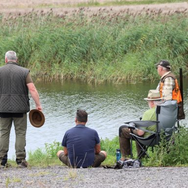 Vier Personen stehen und sitzen am Ufer eines Gewässers, umgeben von Grün.